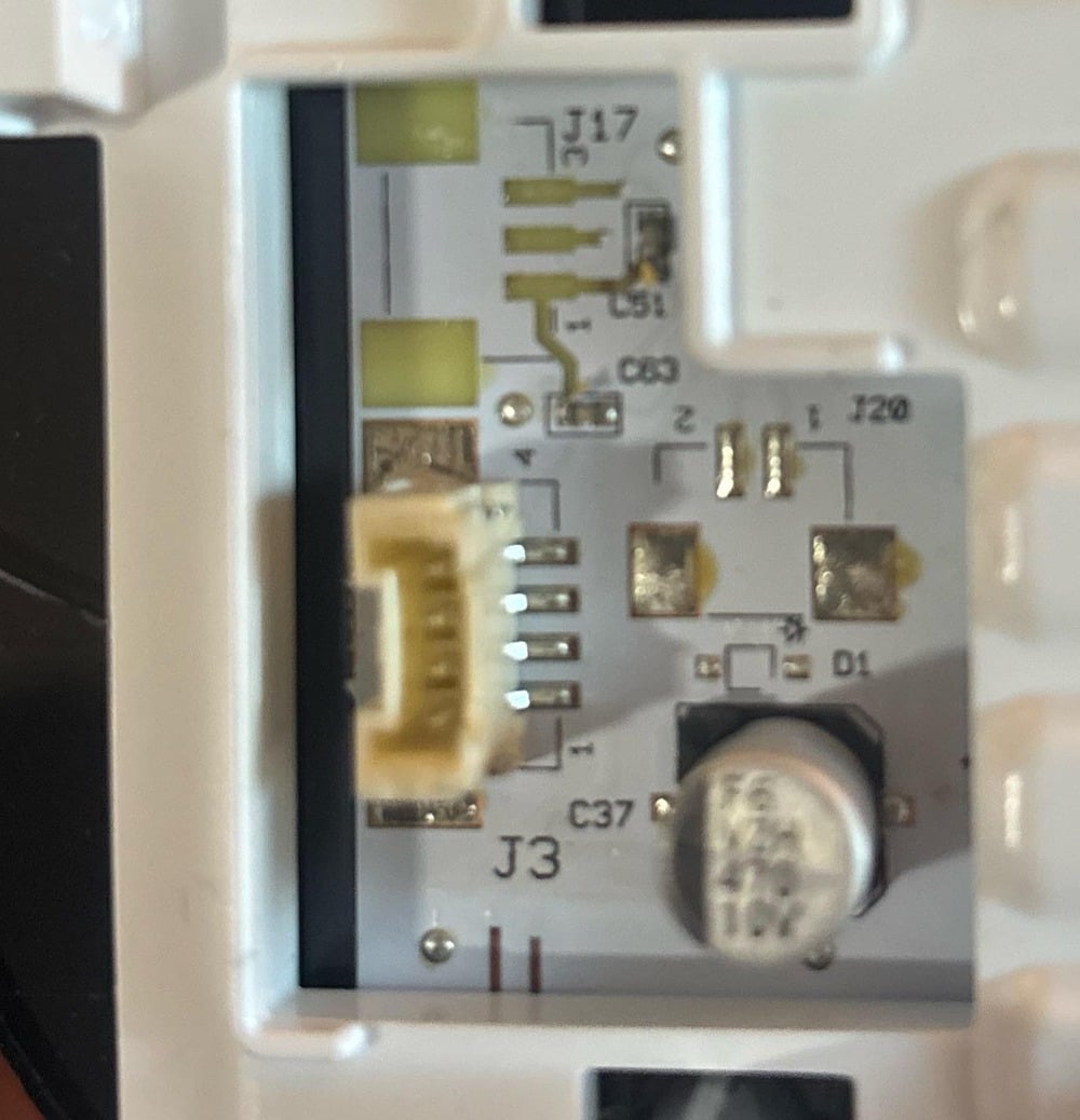 Close-up of a circuit board with visible electronic components, including a white connector, capacitor, and labeled areas such as J3, J17, and C37 mounted on a white plastic surface.