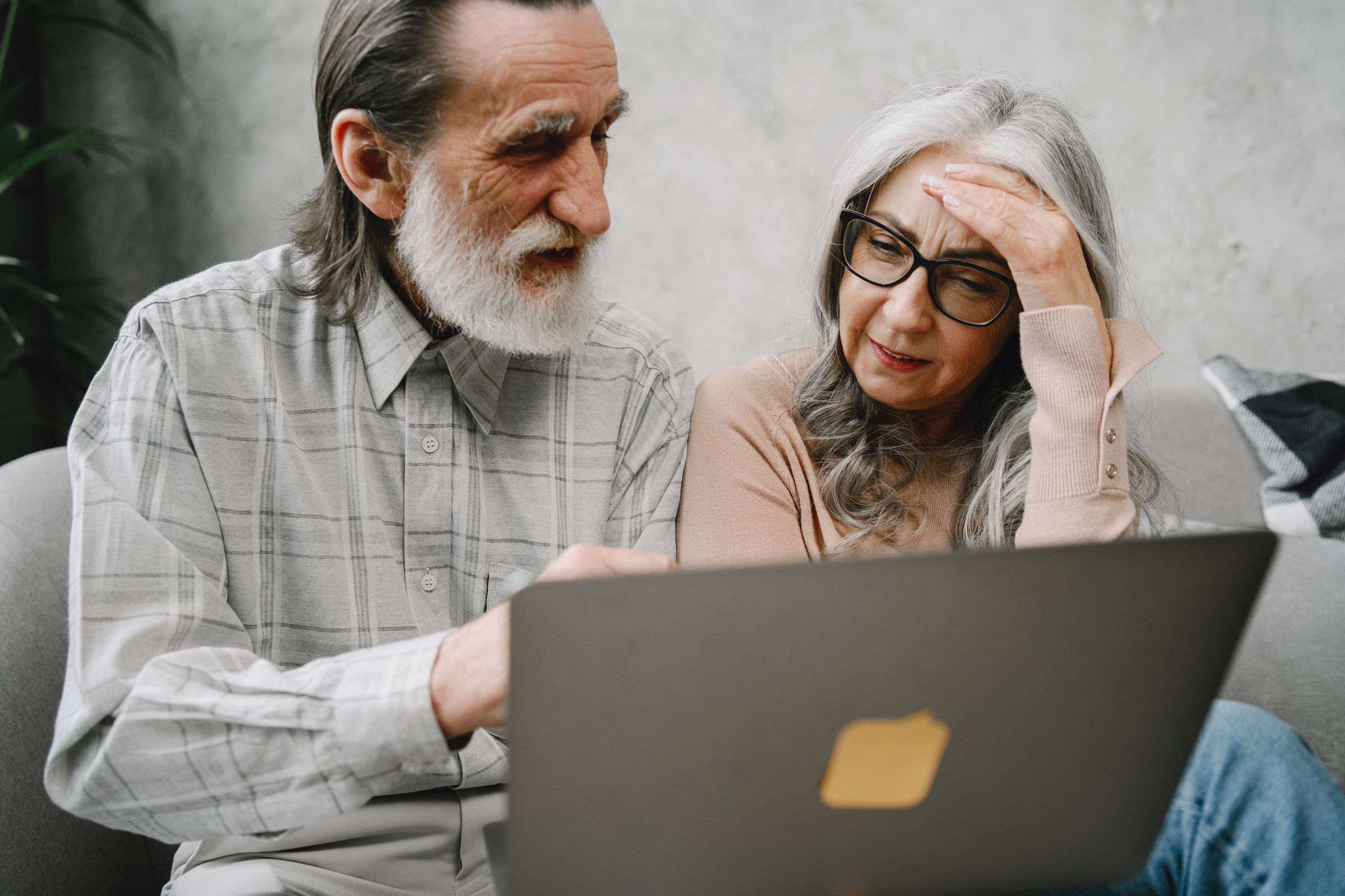 An older man and woman sit closely together on a couch, looking at a laptop screen. The man points at the laptop while the woman rests her hand on her forehead, both appearing engaged.