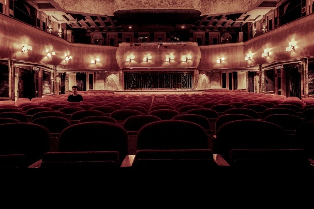 A person sits alone in a large, empty theater filled with rows of dark velvet seats.