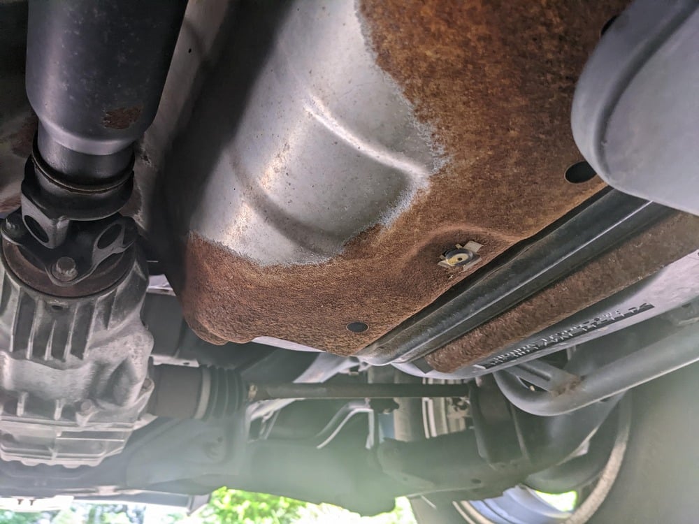 The underside of a vehicle showing significant surface rust on the fuel tank and surrounding metal components.