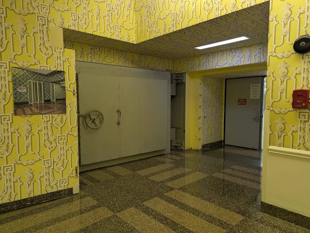 A hallway with yellow patterned wallpaper, a large metal vault door, and a smaller door labeled “Fire Door Keep Closed.”