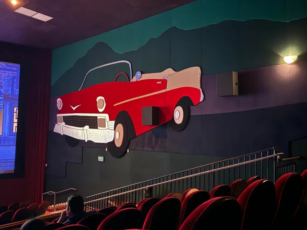 Rows of theater seats facing a wall decorated with a large, stylized cutout of a red vintage convertible car against a painted mountain backdrop.