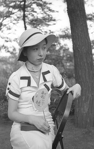 A young woman wearing a sun hat and a collared outfit sits outdoors holding a small round fan.