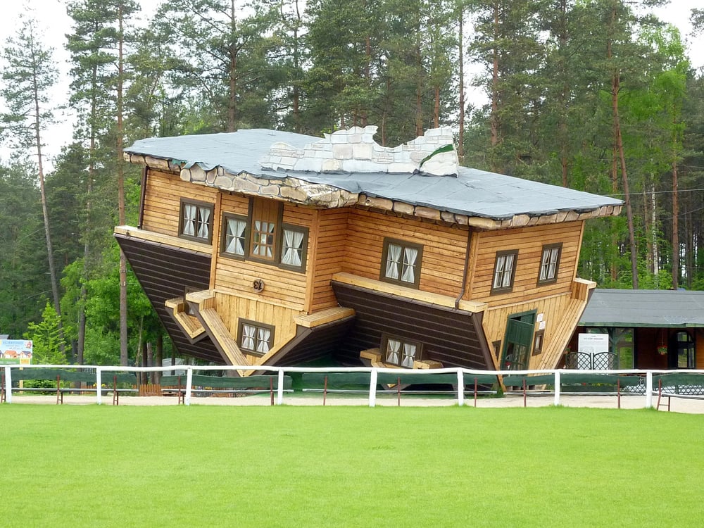 An upside-down wooden house balanced on its roof in a park.