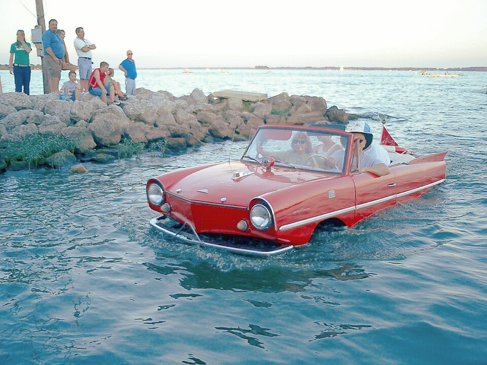 A red amphibious car with passengers drives through the water near a rocky shore where onlookers are gathered.