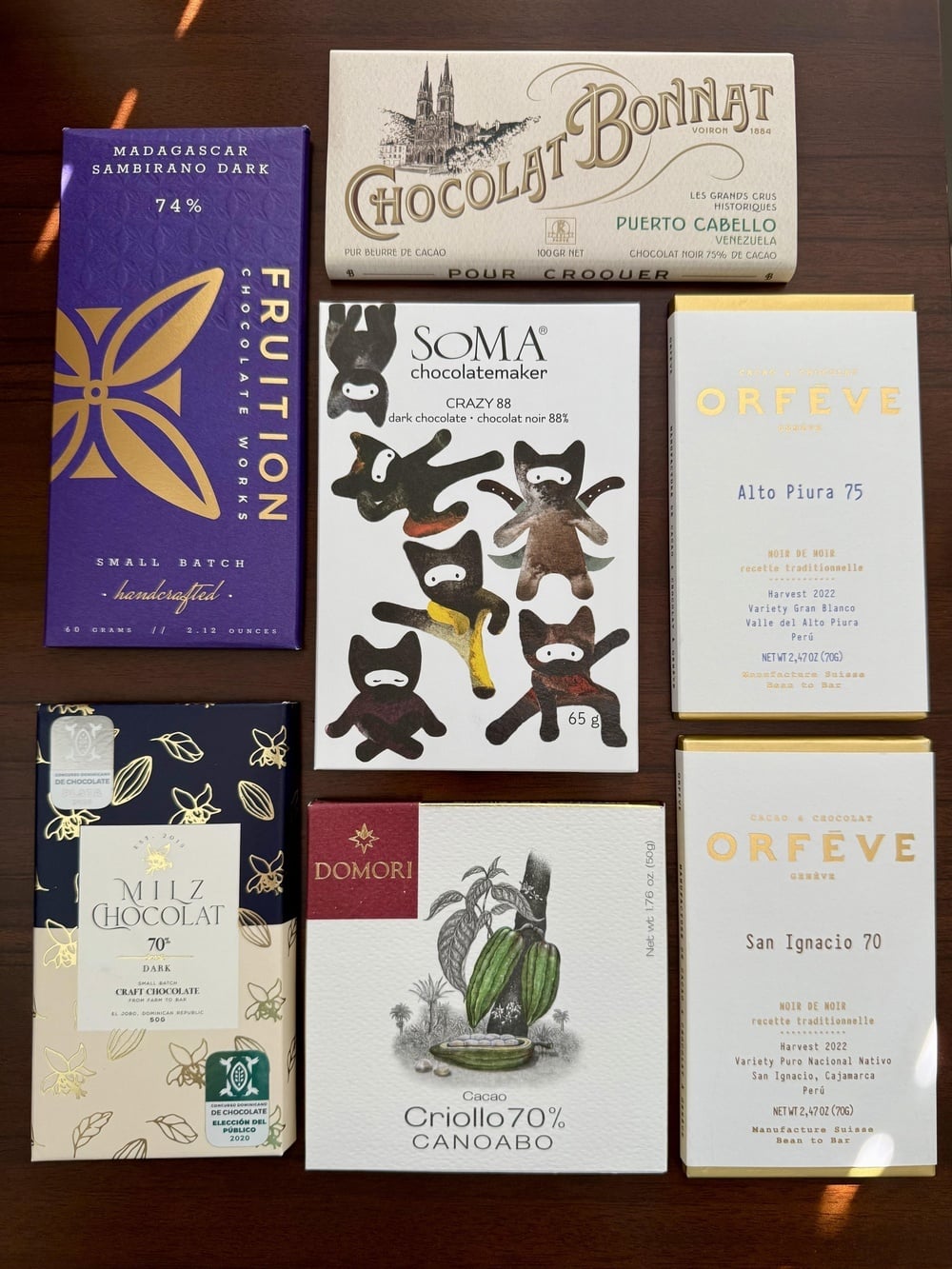 Seven bars of craft chocolate arranged in a grid on a wooden surface, including brands such as Fruition, Bonnat, Soma, Milz, Domori, and Orfève.