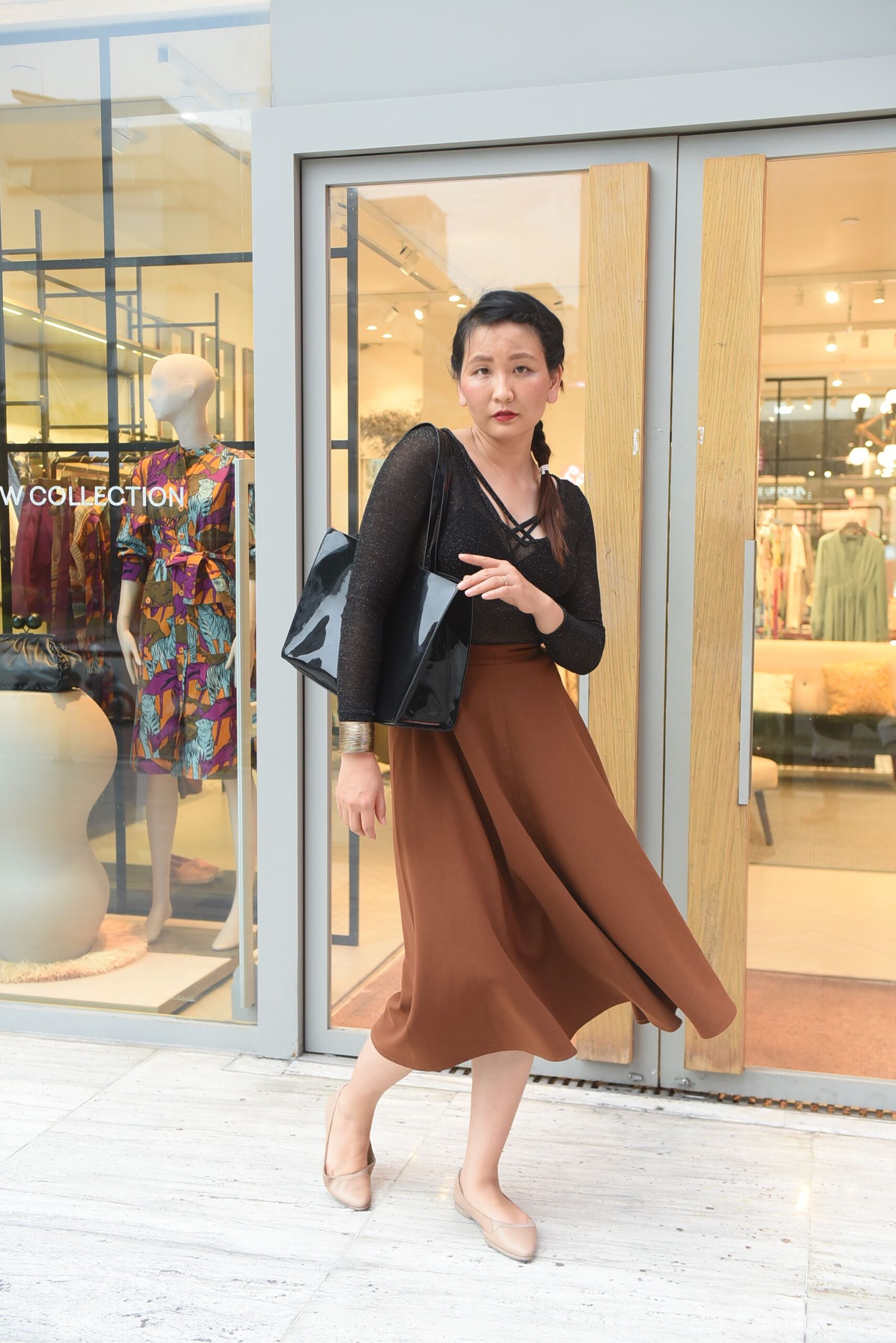 Woman with black handbag and brown midi skirt poses outside a clothing store with mannequins and colorful dresses visible in the window display behind her.