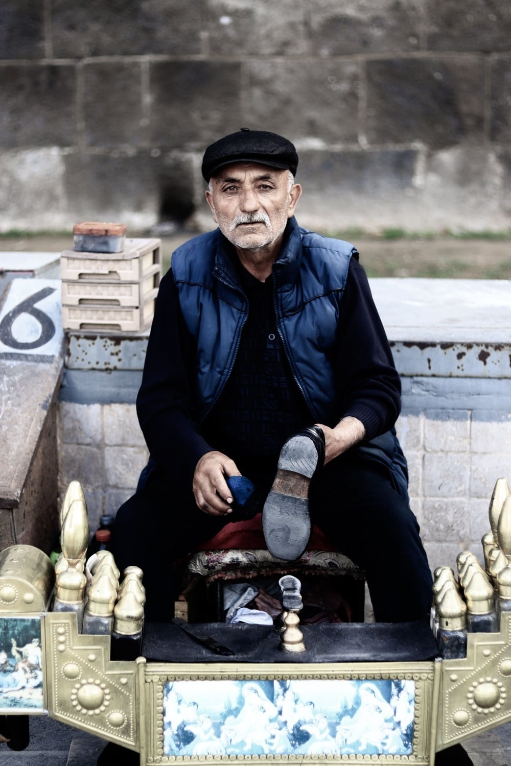 An older man in a cap and vest sits behind a professional shoe-shining kit, holding a shoe.
