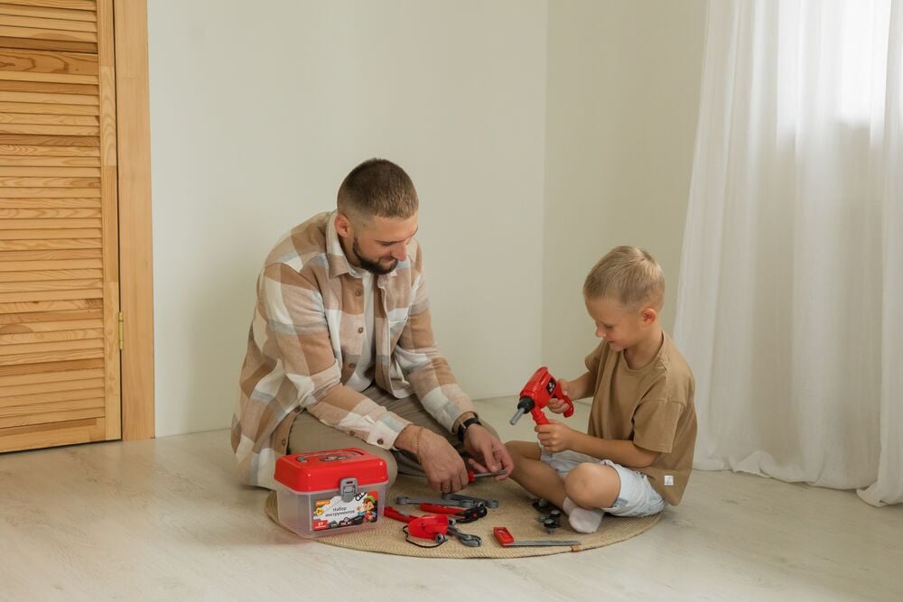 A father and his young son sitting on a rug, playing with a toy tool set and a plastic toolbox.