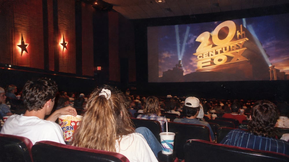 An audience watches the 20th Century Fox logo on a movie theater screen.