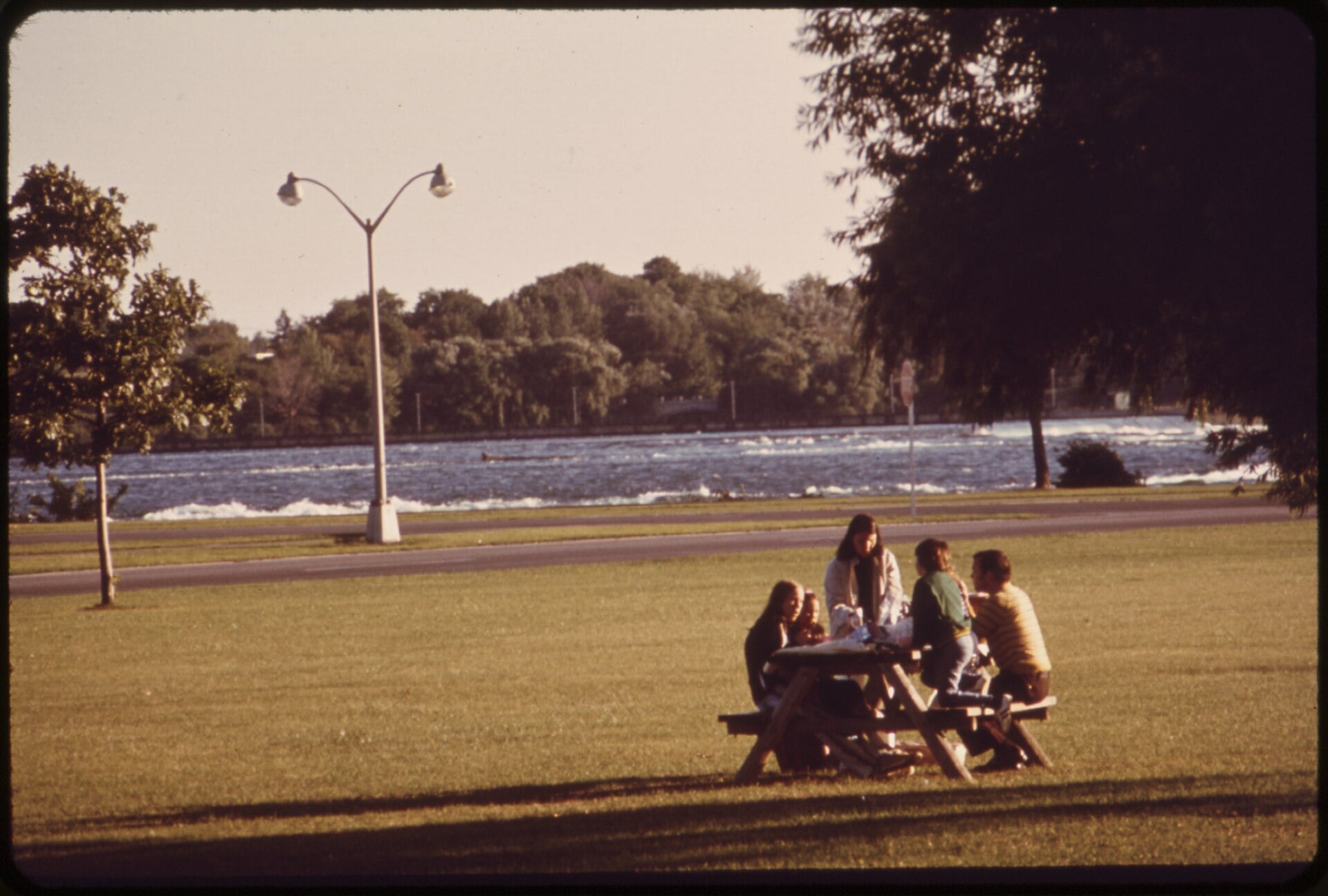 A group of people sits at a picnic table on a grassy lawn beside a river.