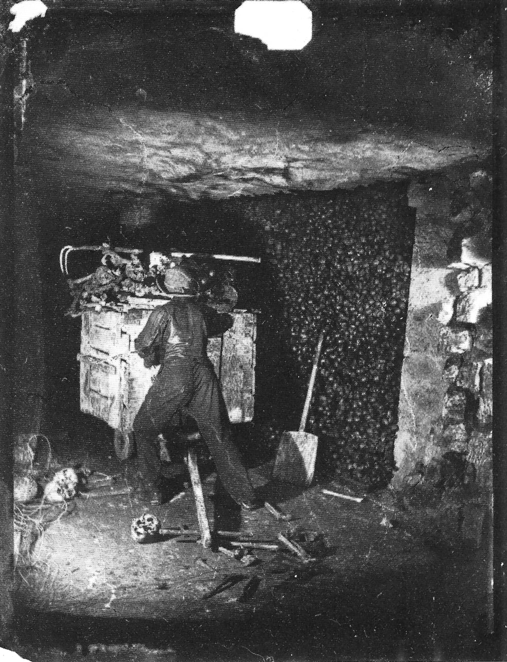 A person in dark clothing pushes a small cart through a tunnel filled with stacks of human skulls and bones.