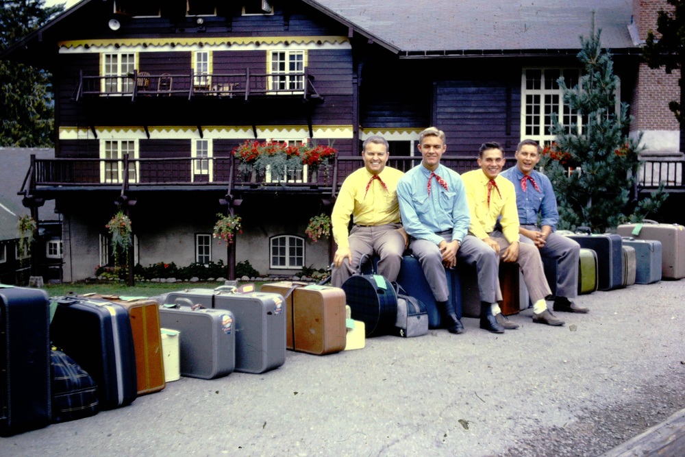 Four men in matching shirts and red neckerchiefs sit on suitcases outside a dark wooden lodge.