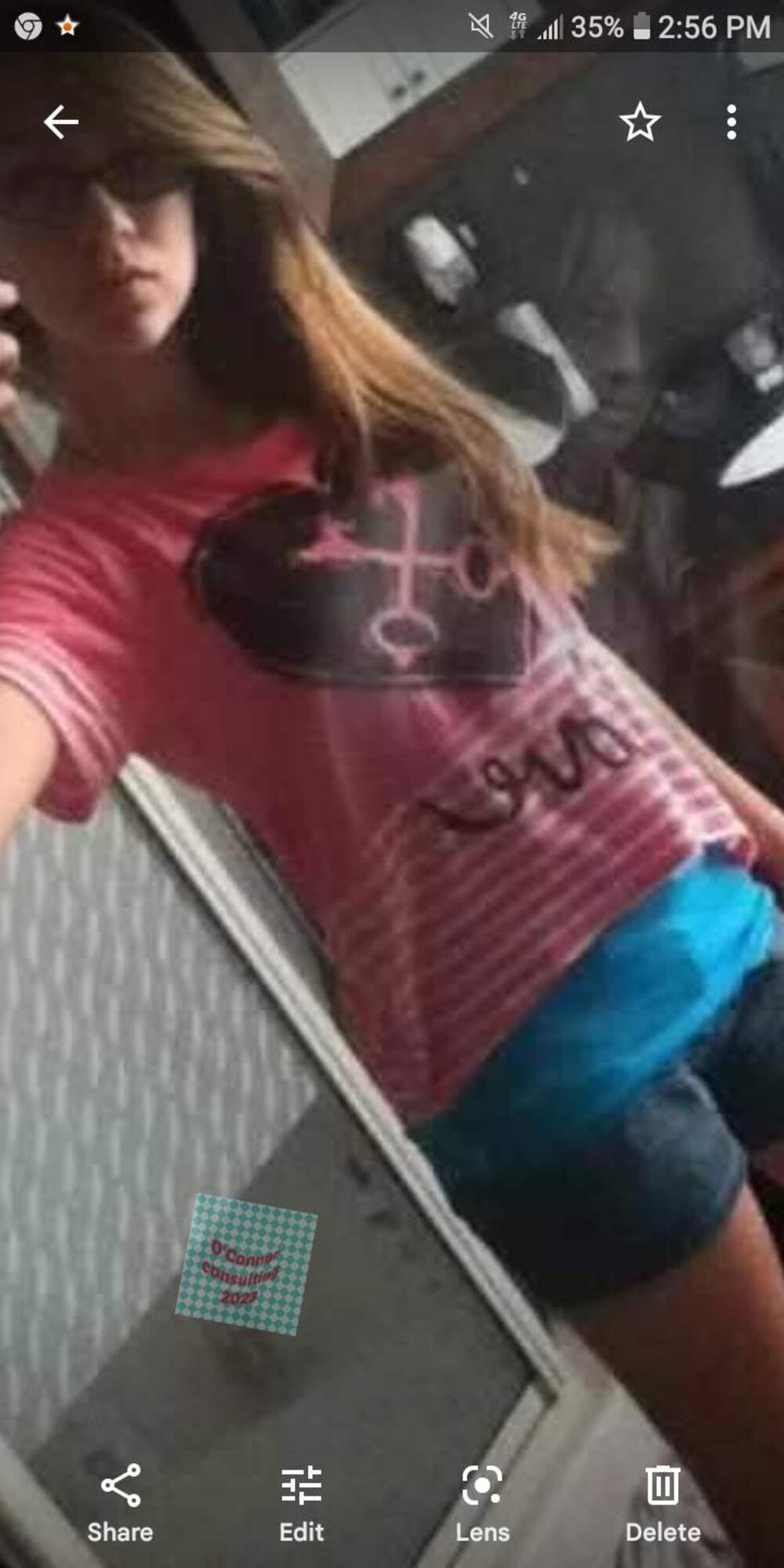 A person wearing a pink striped shirt with a black heart graphic and blue shorts taking a selfie in a mirror.