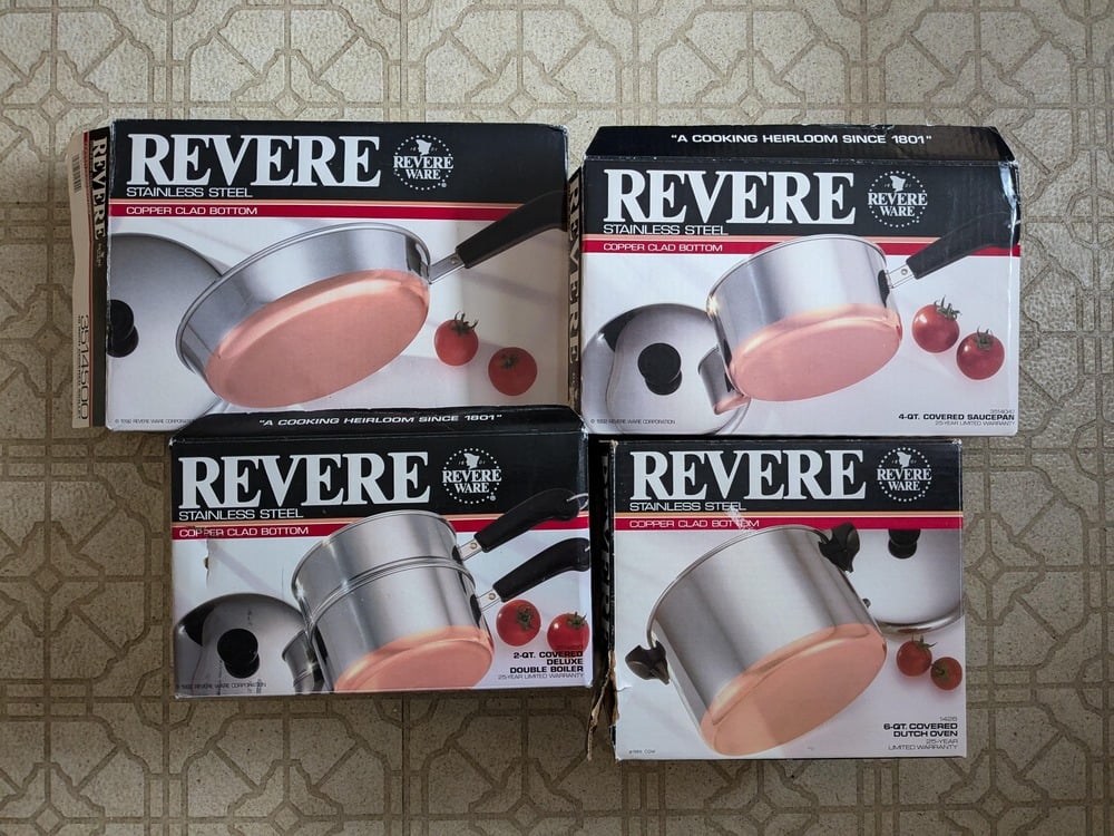 Four Revere stainless steel cookware boxes with copper-clad bottoms are arranged on a patterned floor. Each box features images of pots or pans and tomatoes, and displays the Revere brand name prominently.