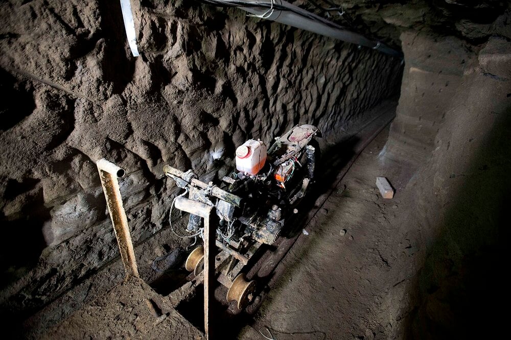 A stripped motorcycle frame mounted on small wheels sits on a track inside a narrow, dark dirt tunnel.