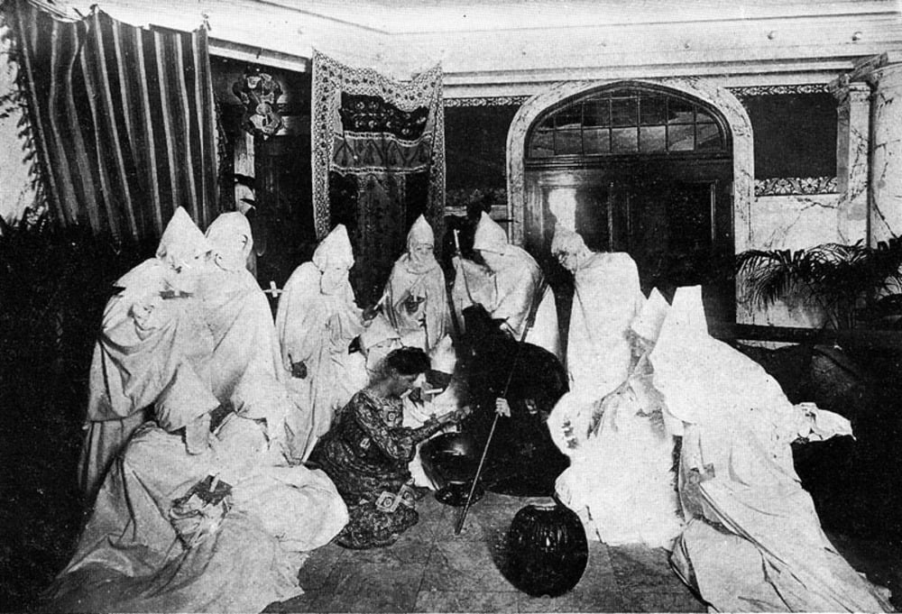 A group of people wearing Ku Klux Klan robes and pointed hoods gather around a figure in patterned clothing kneeling on the floor.