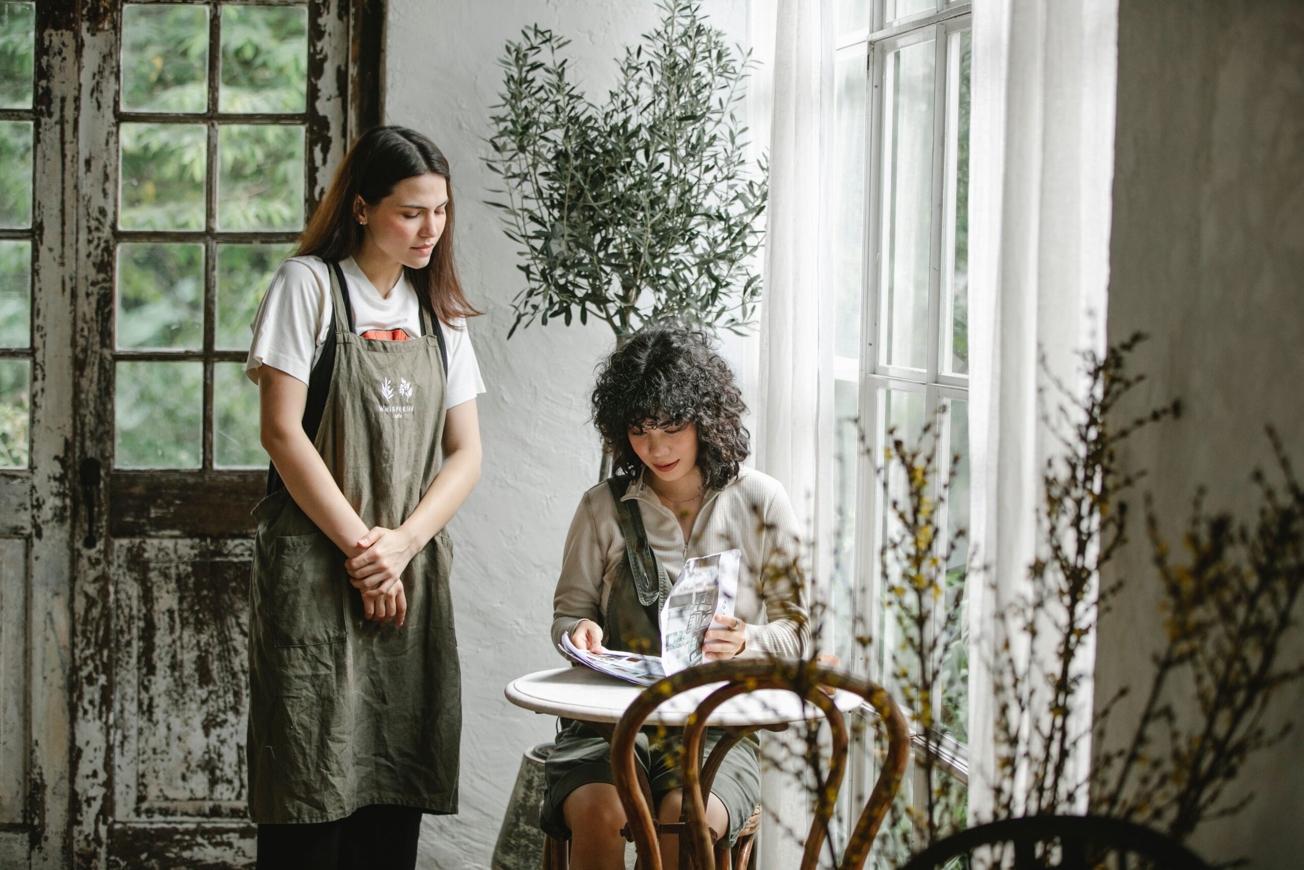Two women in a rustic cafe, one standing in an apron beside the other who is seated at a small table by a window, reading a magazine. Natural light streams in, highlighting green plants and a wooden chair.