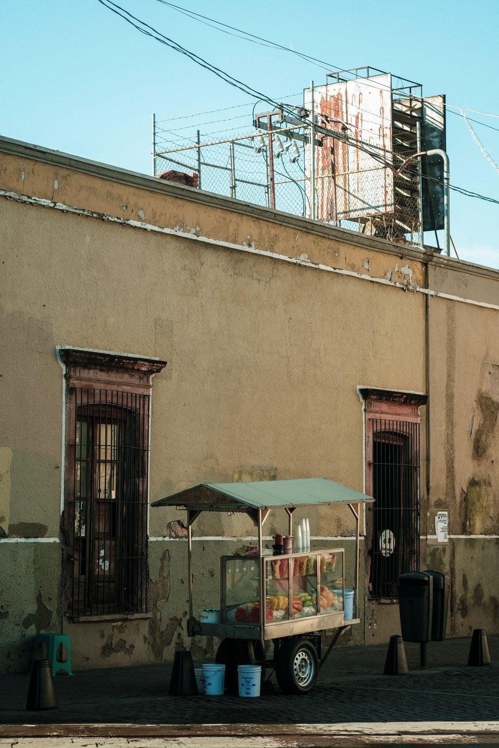 A mobile street food cart with a glass display case stands on a cobblestone street in front of a weathered, two-story building.