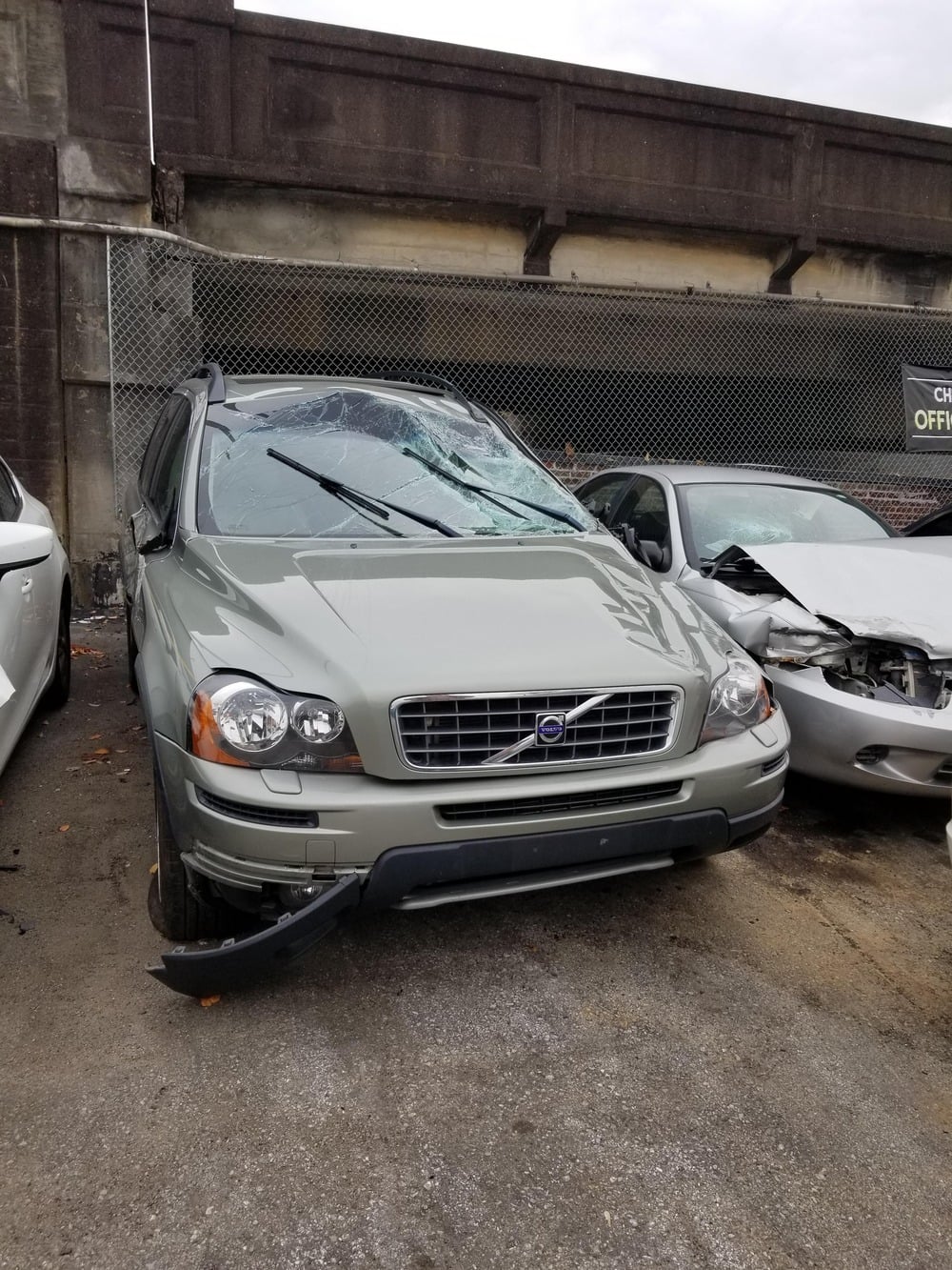 A light green Volvo SUV with a shattered windshield and a detached front bumper parked in a lot.