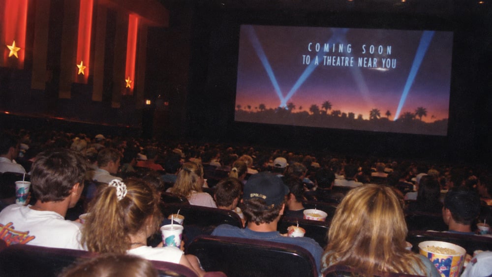 An audience sits in a movie theater facing a large screen displaying the words “Coming soon to a theatre near you” over a silhouette of palm trees and spotlights.