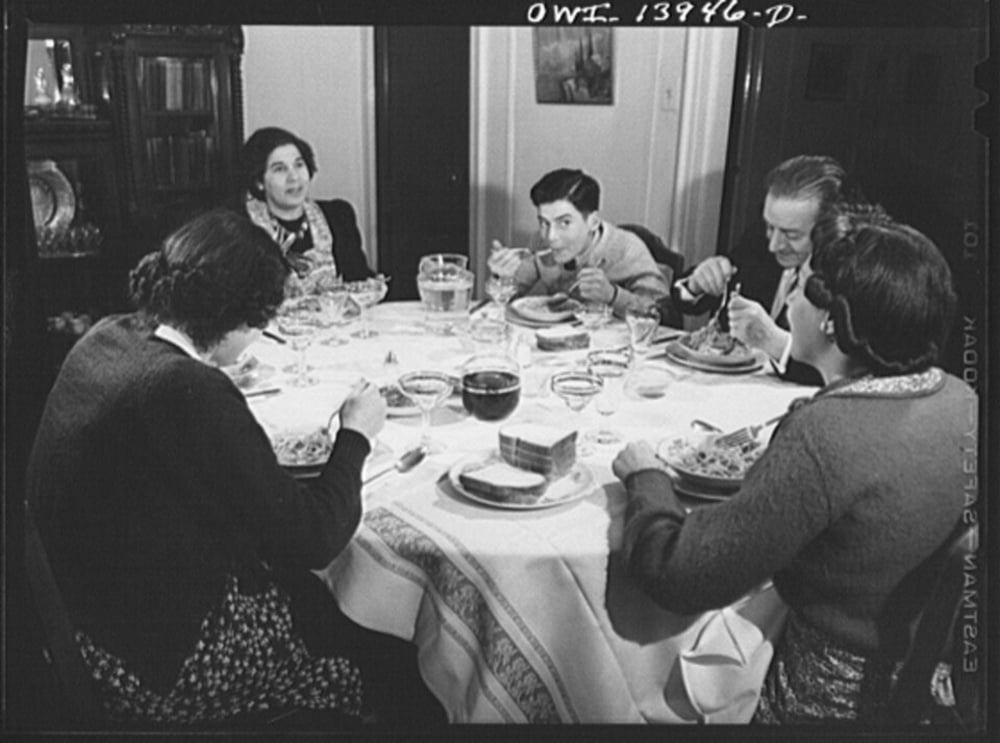 Five people sit around a dining table set with plates of food, glasses, and a pitcher.