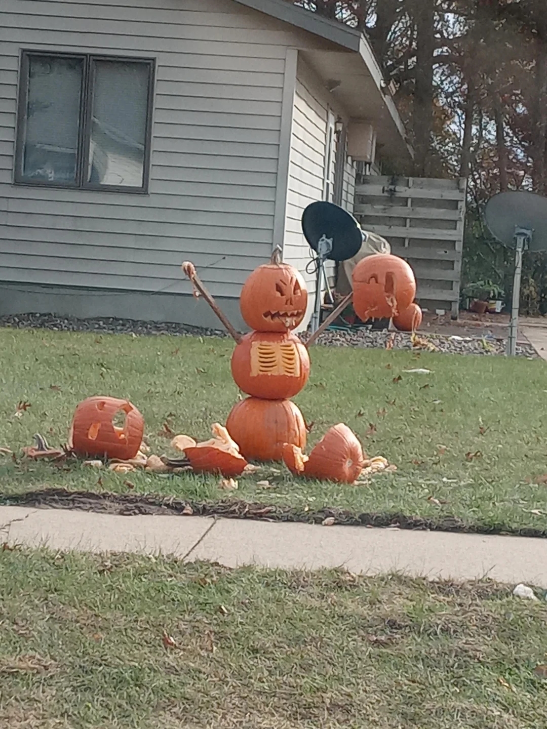 A snowman made of three stacked pumpkins in a front yard, with carved faces, a ribcage design, and sticks for arms.