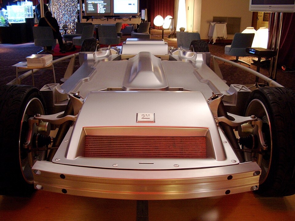 A silver General Motors vehicle chassis with exposed suspension and a prominent front radiator, displayed in an indoor room.