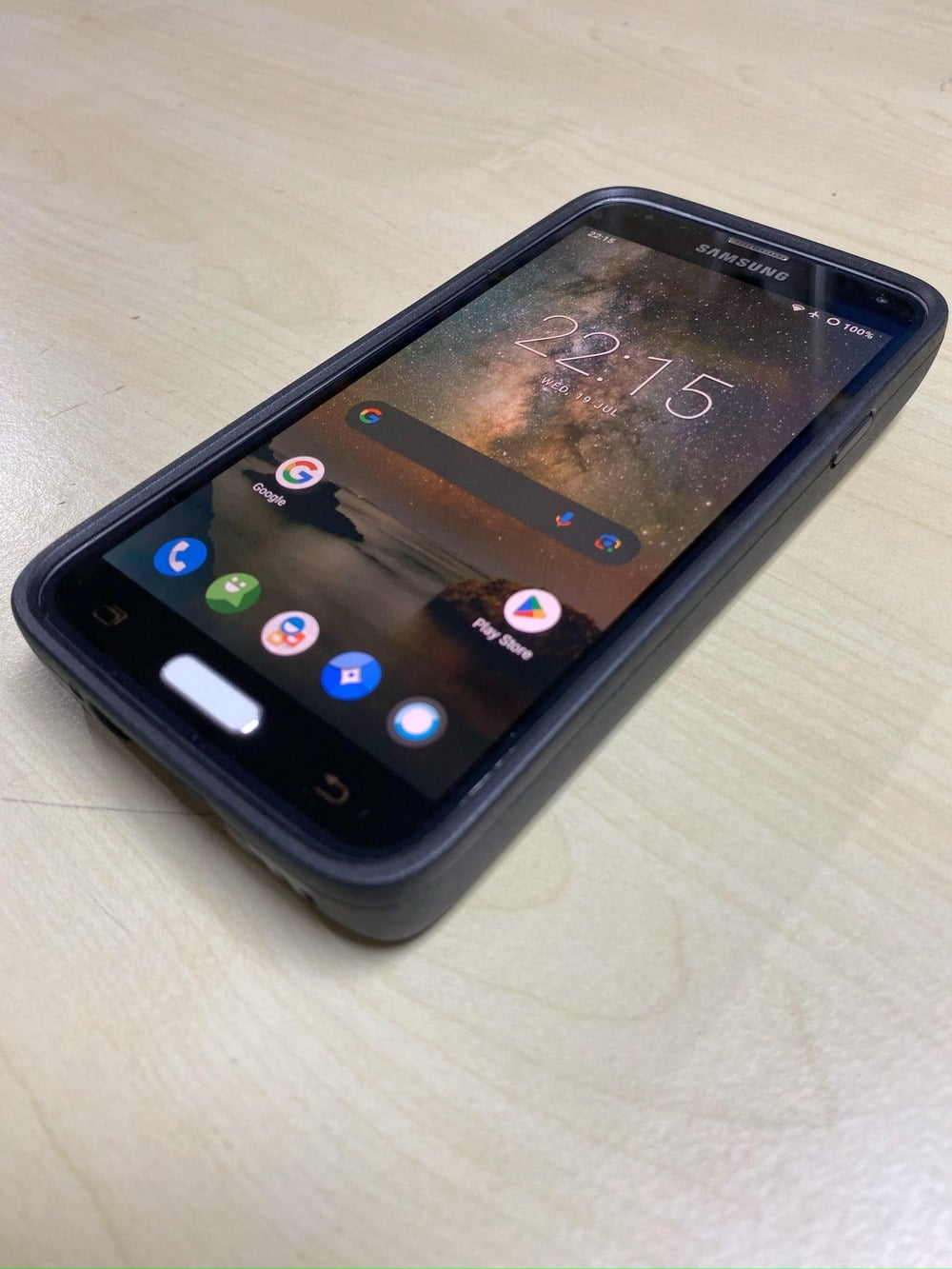 A Samsung smartphone with a case rests on a light-colored surface, displaying the home screen with app icons and a background image of a starry night sky. The time shown is 22:15.