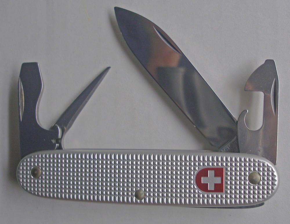 A silver Swiss Army knife with a large blade, can opener, bottle opener, and reamer unfolded, lying on a white surface.