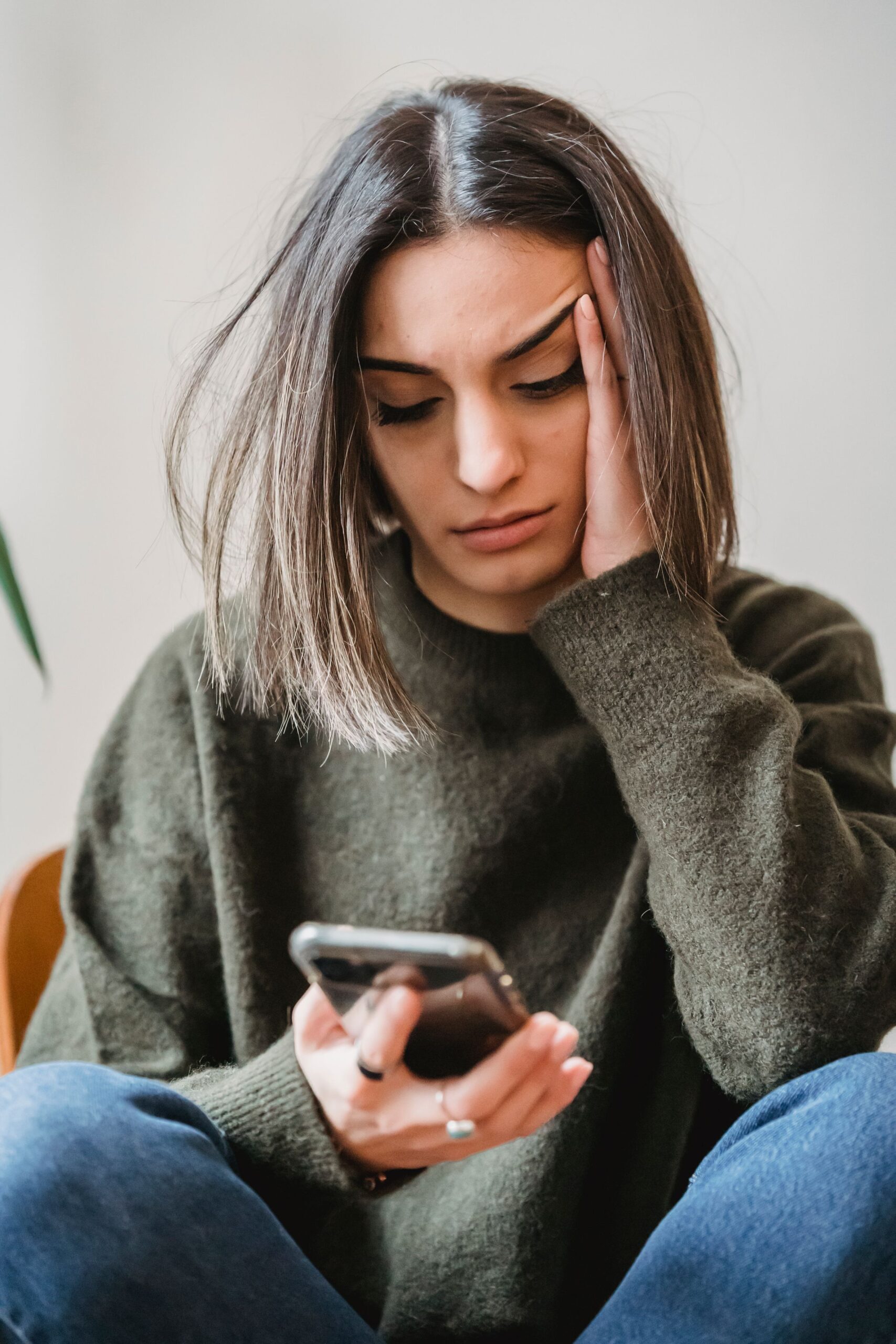 A woman with shoulder-length brown hair looks stressed as she holds her phone in one hand and rests her head on the other, sitting indoors in casual clothes.