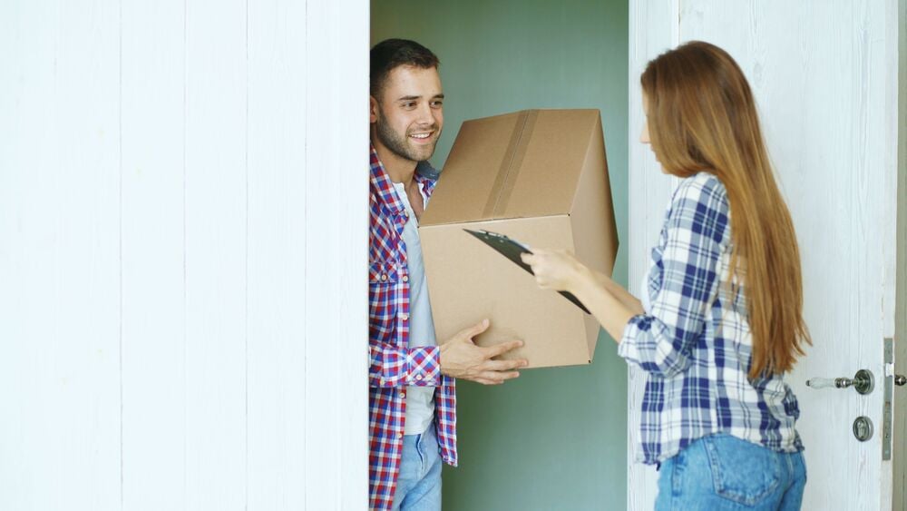 A woman hands a cardboard box to a smiling man standing in a doorway.