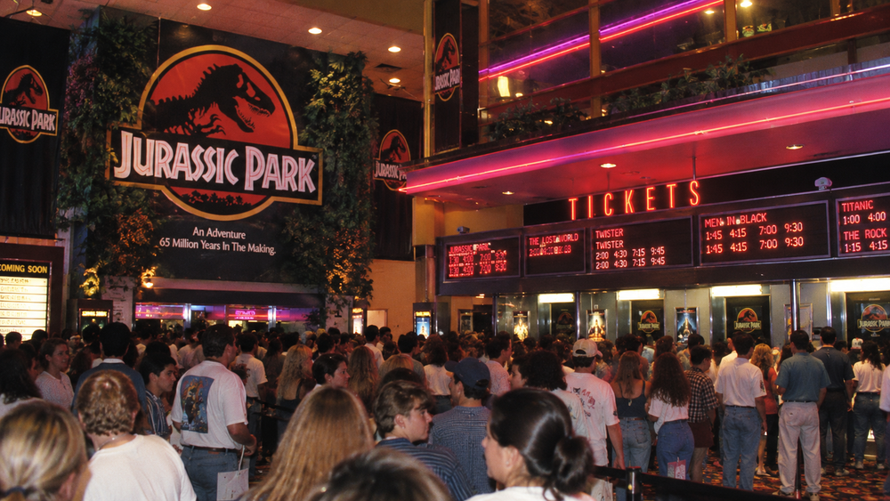 A large crowd of people standing in a movie theater lobby decorated with Jurassic Park signage and a ticket booth with digital showtime displays.