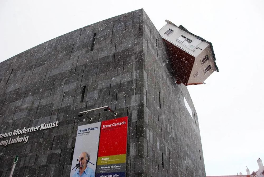 A small house perched at an angle on the corner of a dark, stone-clad museum building during a snowstorm.