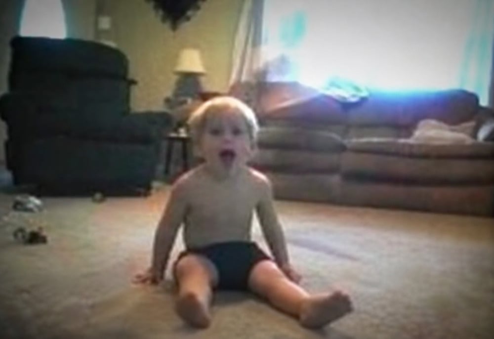 A toddler sits on a carpeted floor with their mouth open.