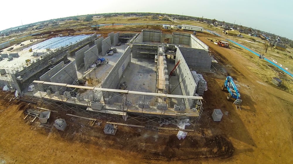 Aerial view of a building under construction, featuring concrete walls, scaffolding, and a blue aerial lift on a dirt construction site.