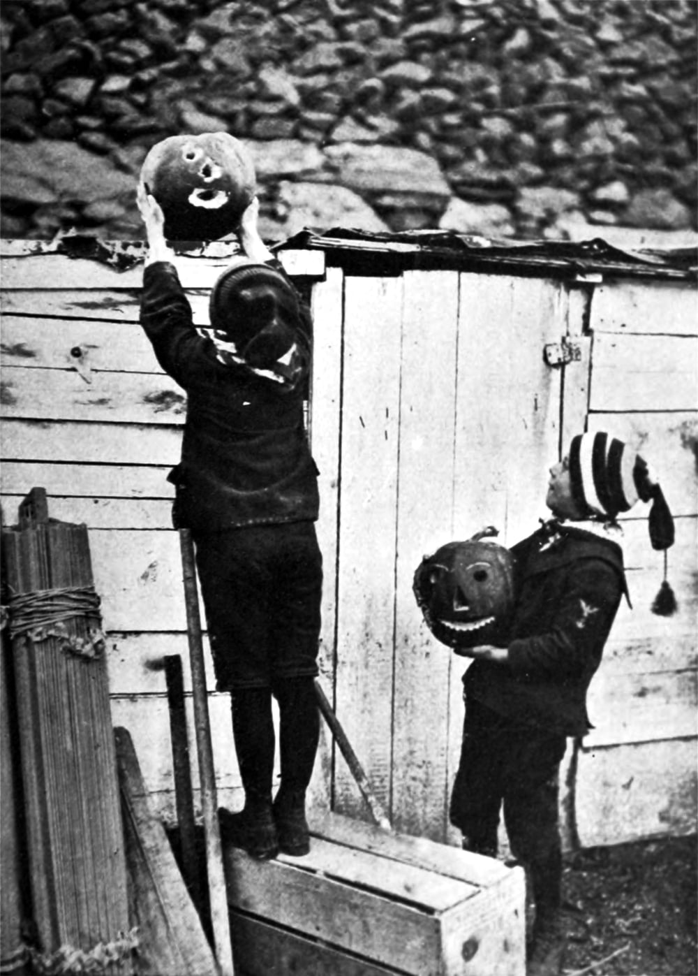 Two children in hats and dark clothing holding carved jack-o’-lanterns outdoors.