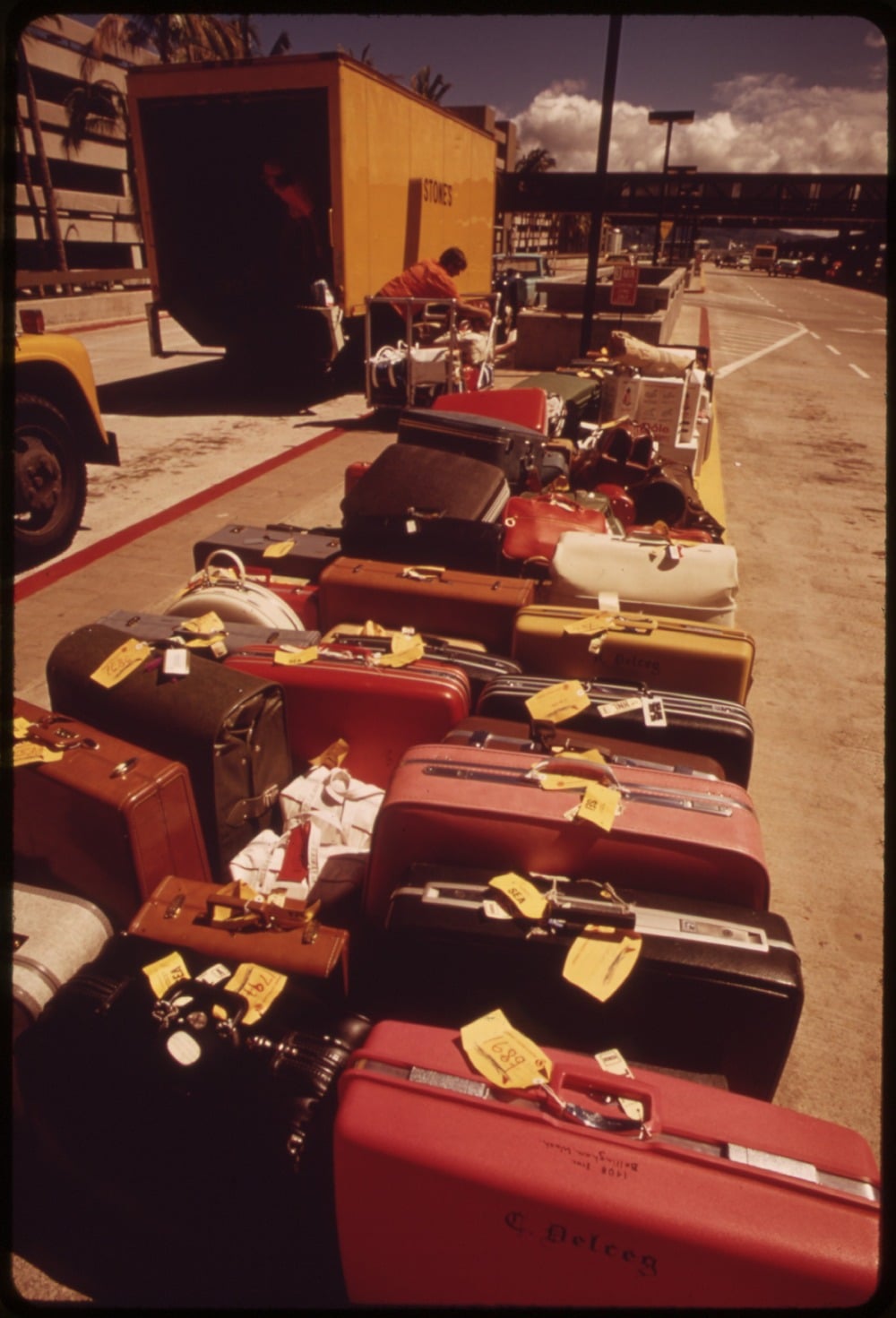 Rows of tagged suitcases and bags line a curb outside, with a yellow transport truck in the background under a partly cloudy sky.
