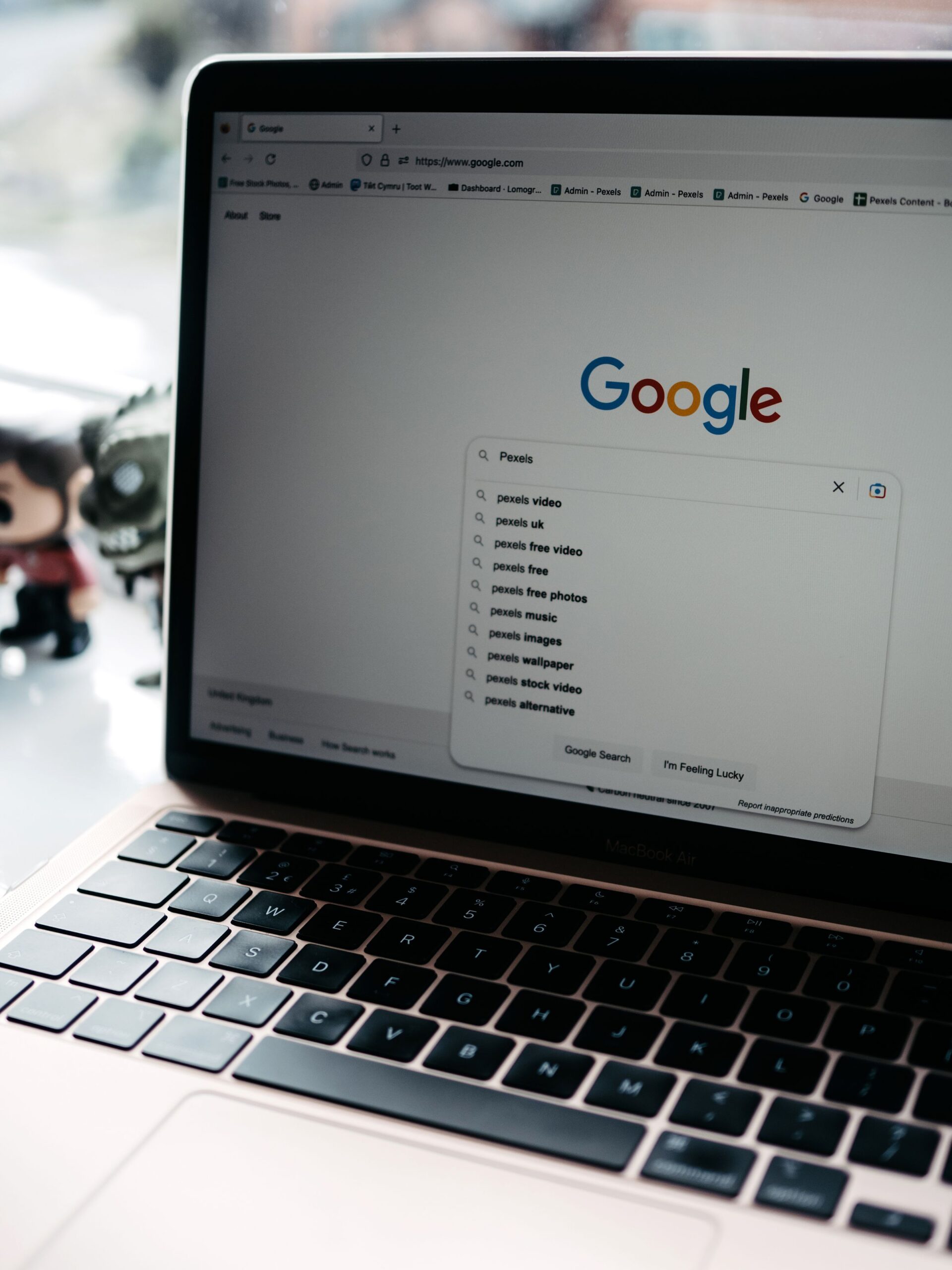 A laptop displays the Google homepage with “Pexels” typed in the search bar and related suggestions. A blurred figurine and window are visible in the background.