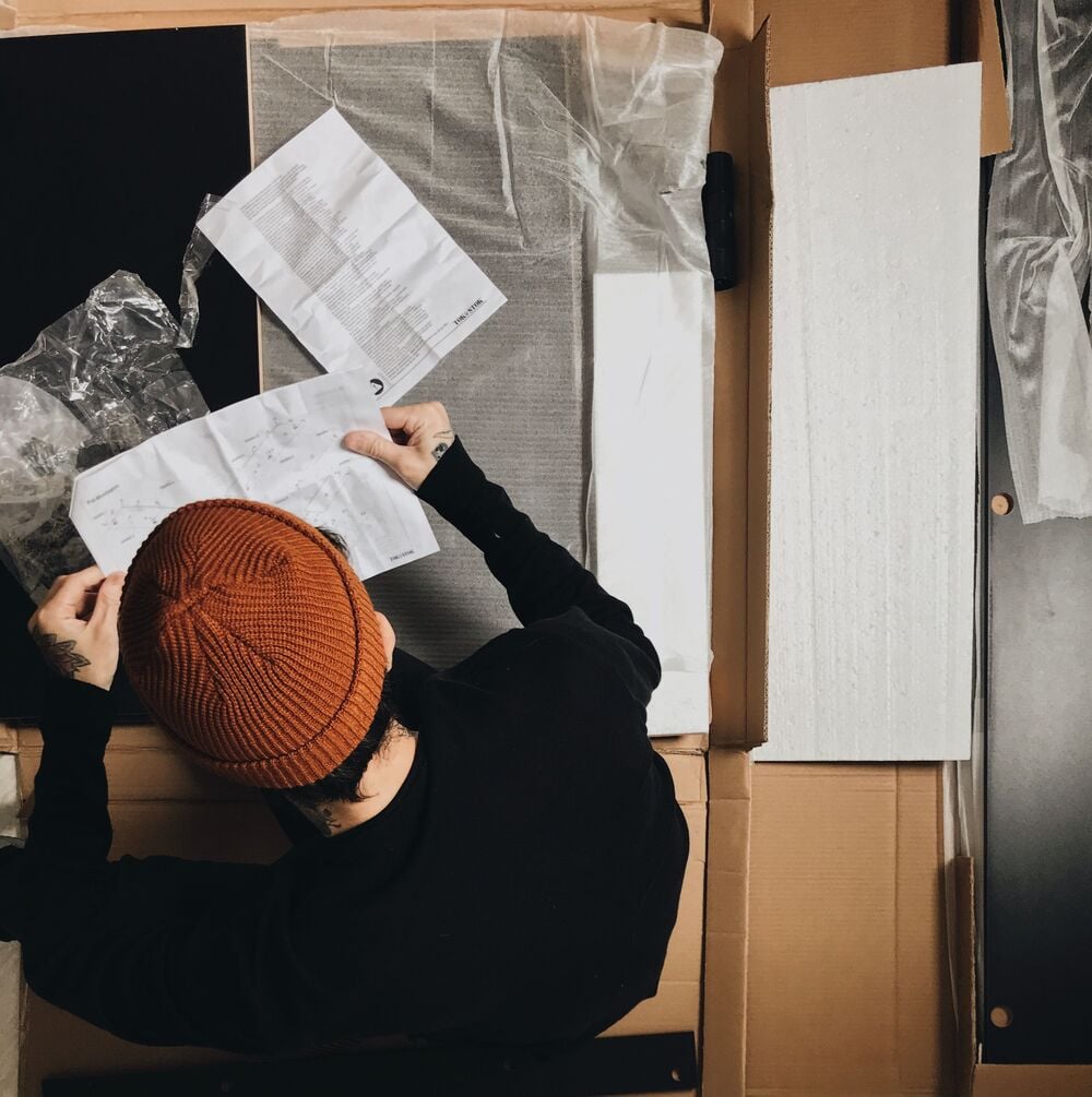 A person reading assembly instructions over an open cardboard box containing furniture parts.