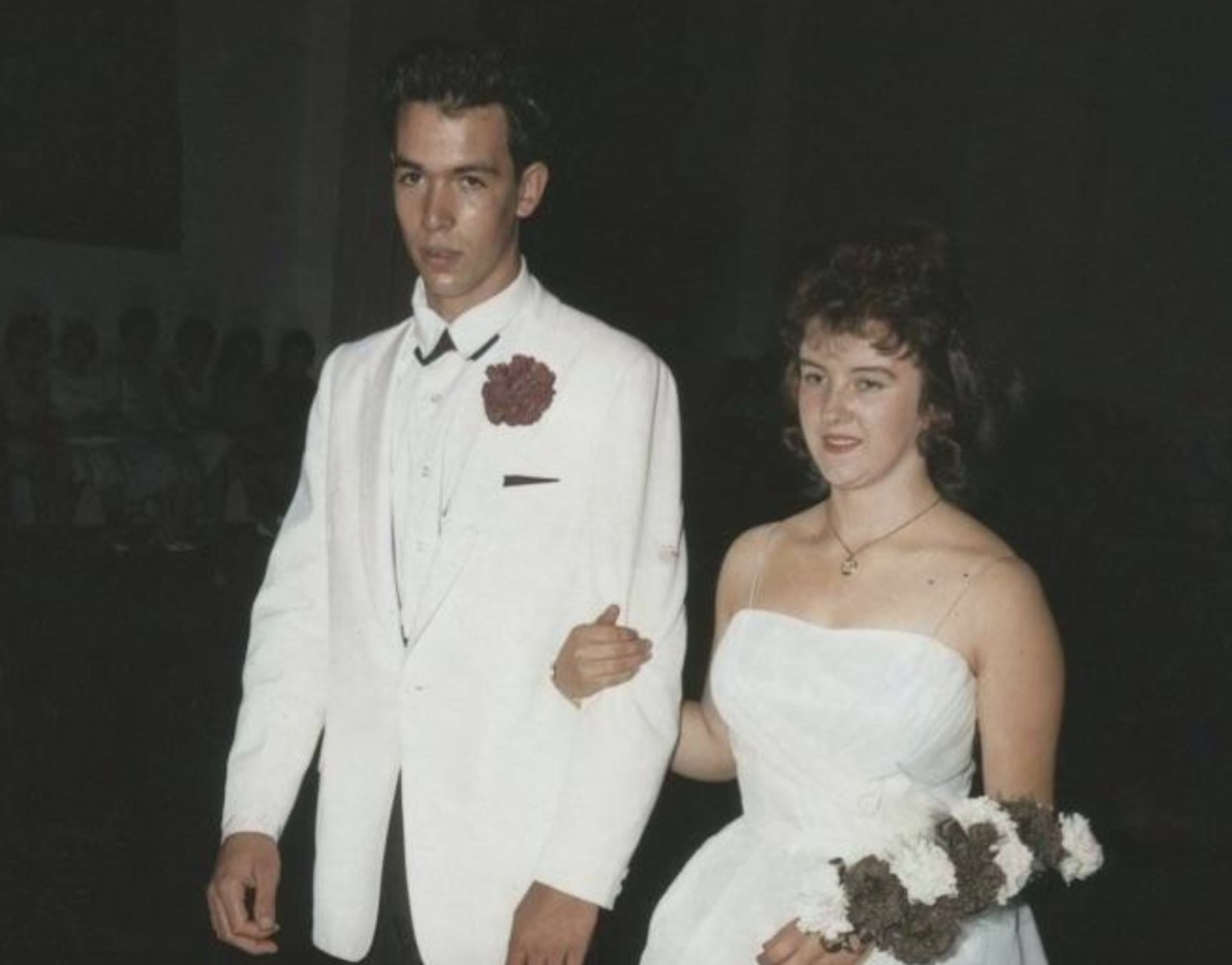 A young man in a white tuxedo with a dark boutonniere walks with a young woman in a white strapless gown holding a floral bouquet.