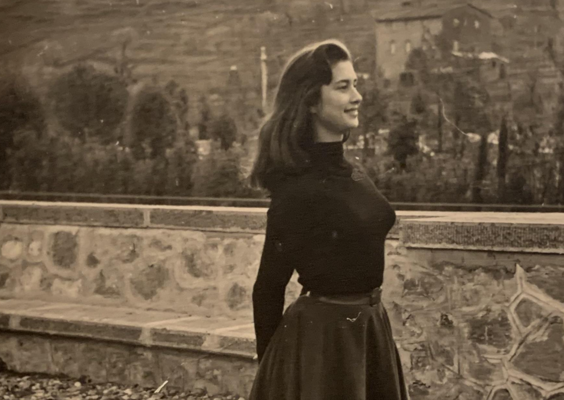 A sepia-toned vintage photograph of a woman in a long-sleeved shirt and skirt standing profile against a stone wall.