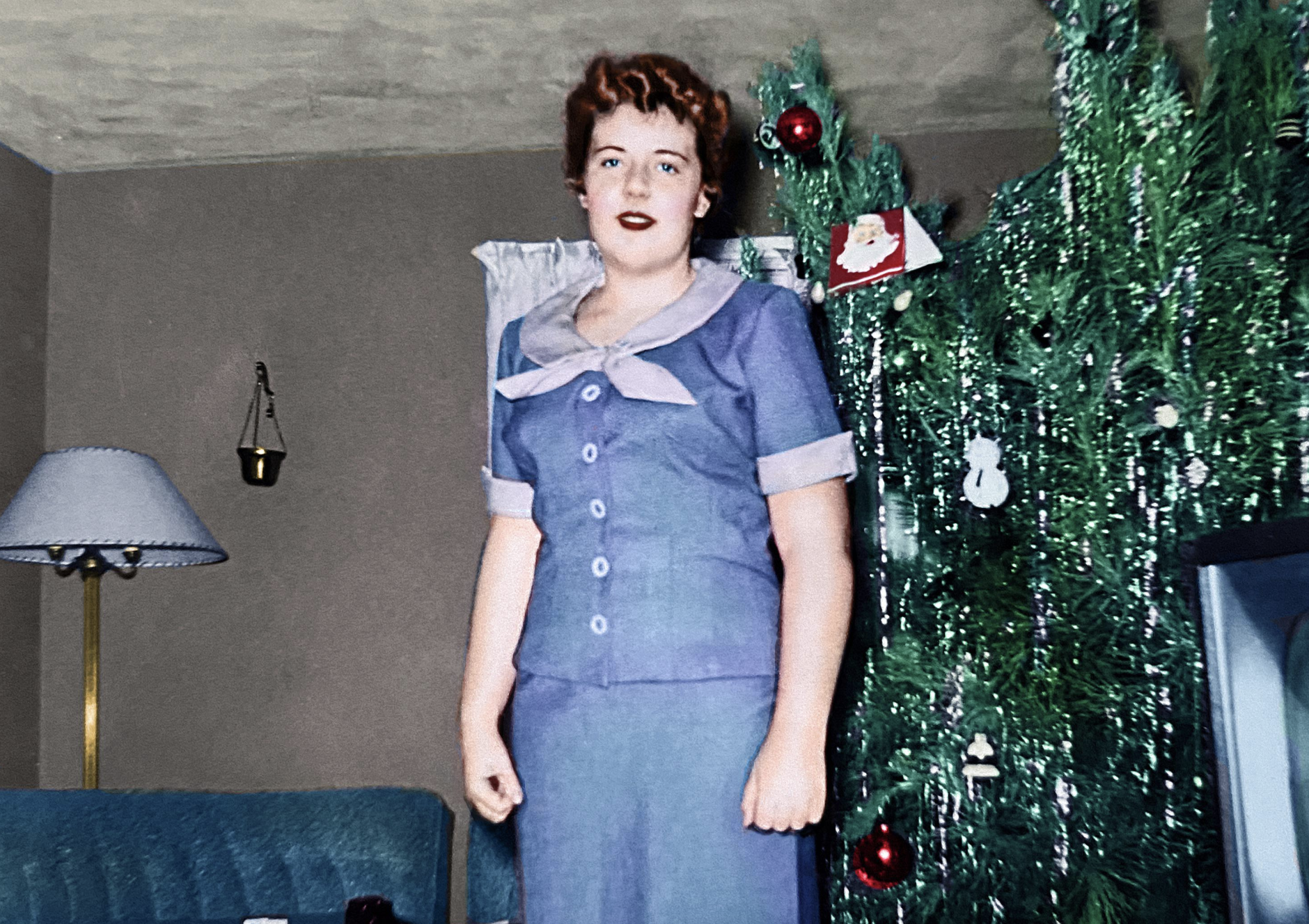 A woman with short auburn hair wearing a blue skirt suit stands next to a decorated Christmas tree.