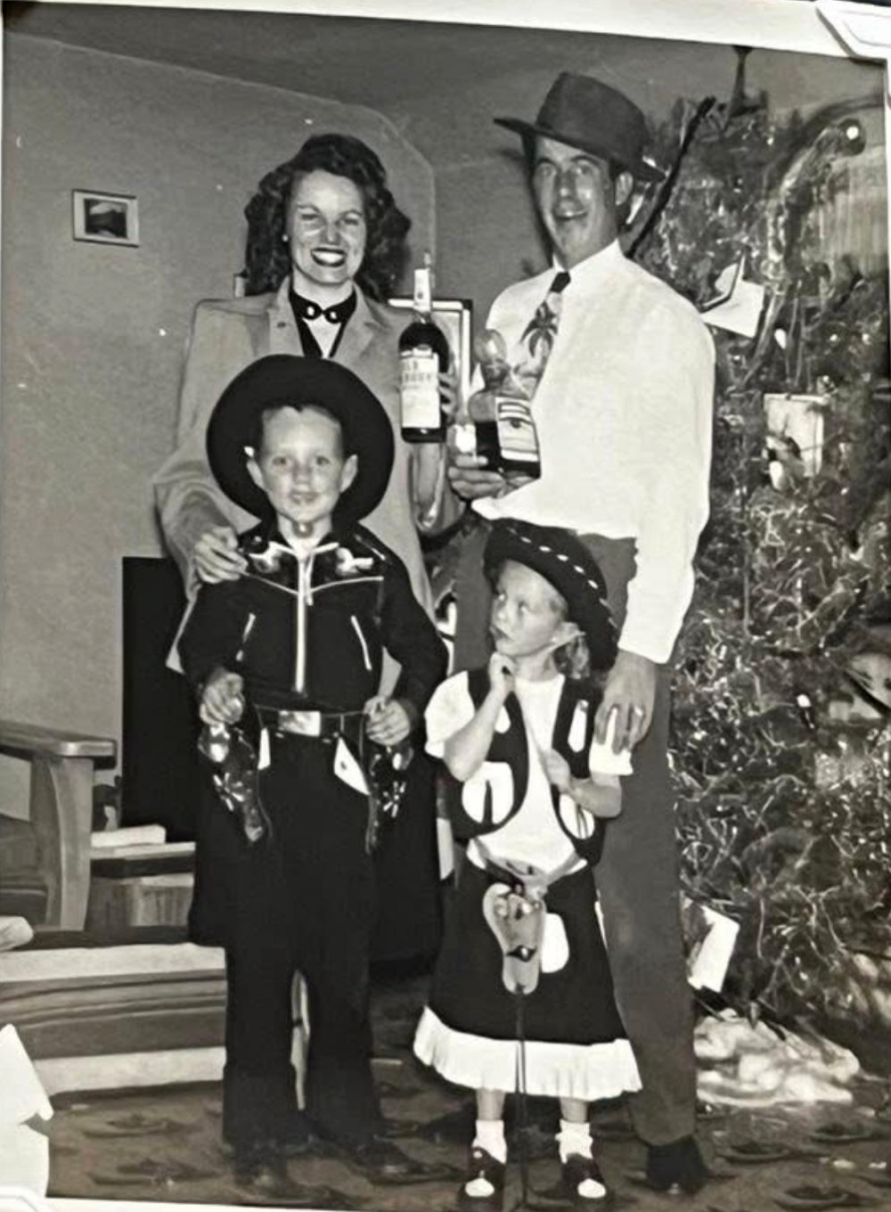 A black-and-white family portrait featuring a woman and man standing behind a boy and girl dressed in western-style cowboy and cowgirl costumes in front of a Christmas tree.