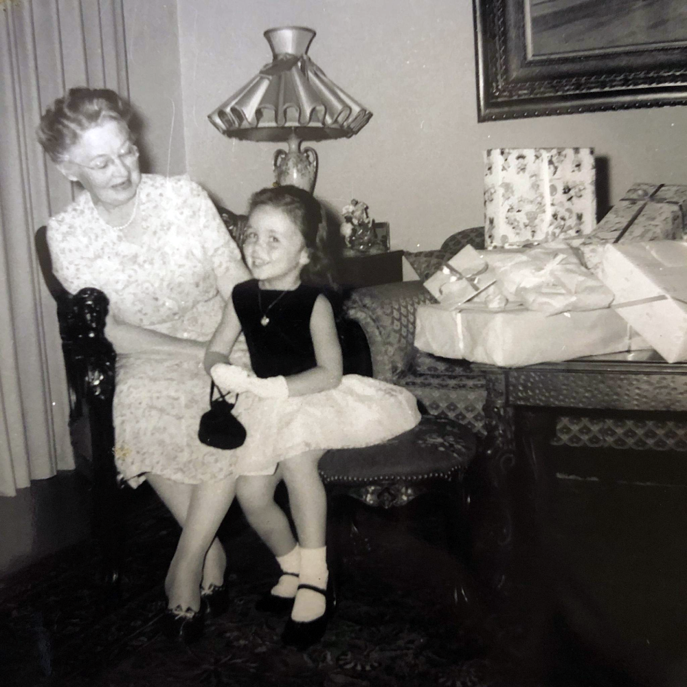 An older woman and a young girl in a dress and gloves sit together on a small sofa next to a table with wrapped gifts.