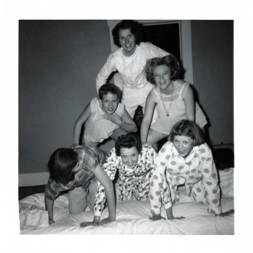 Five young women in pajamas form a human pyramid on a bed.