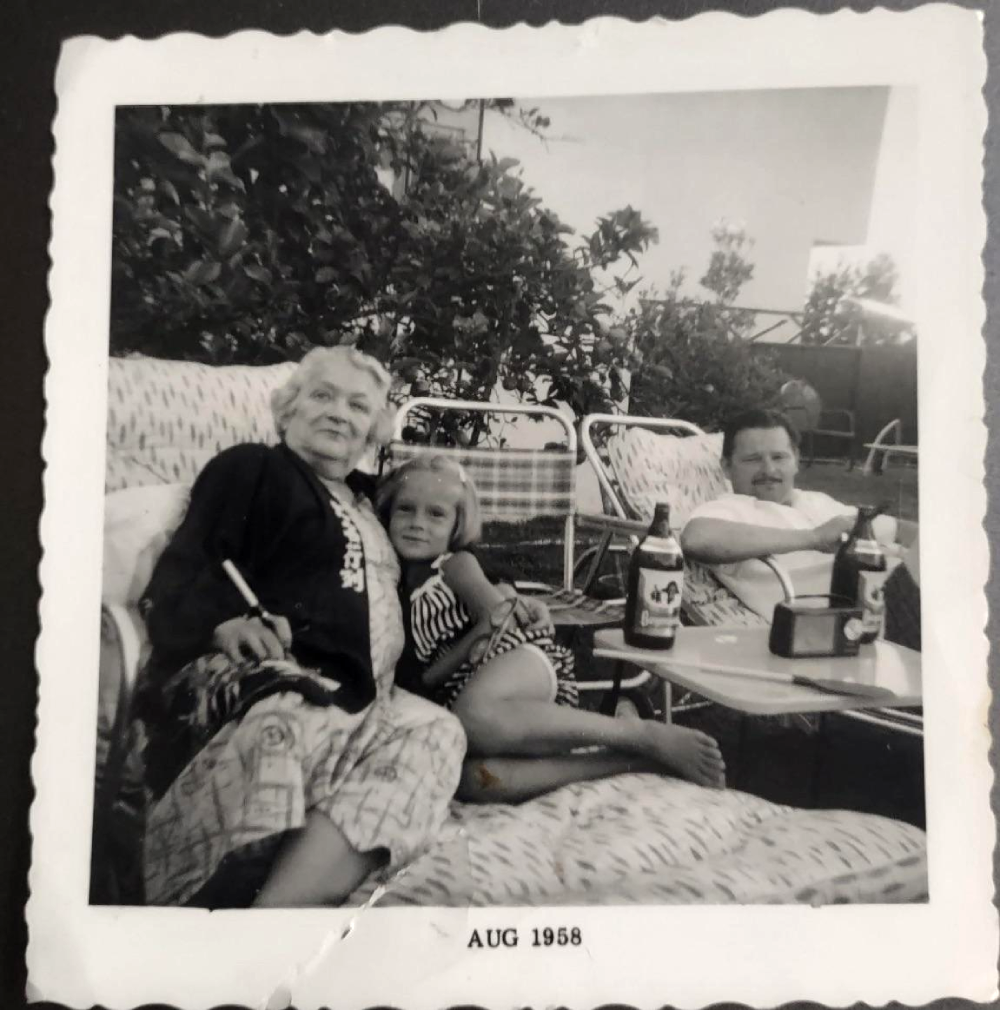 Black and white photo of an older woman, a young girl, and a man sitting on lawn chairs outdoors, dated August 1958.