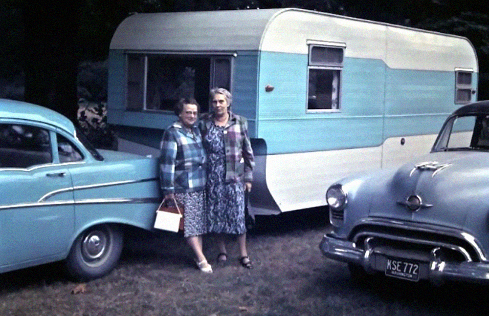 Two women standing between a vintage travel trailer and two parked cars.