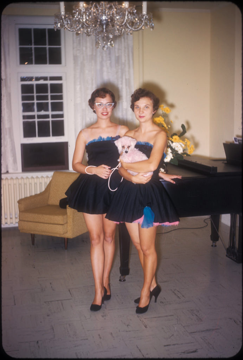 Two women in matching black cocktail dresses pose by a grand piano, one holding a small, light-colored dog.