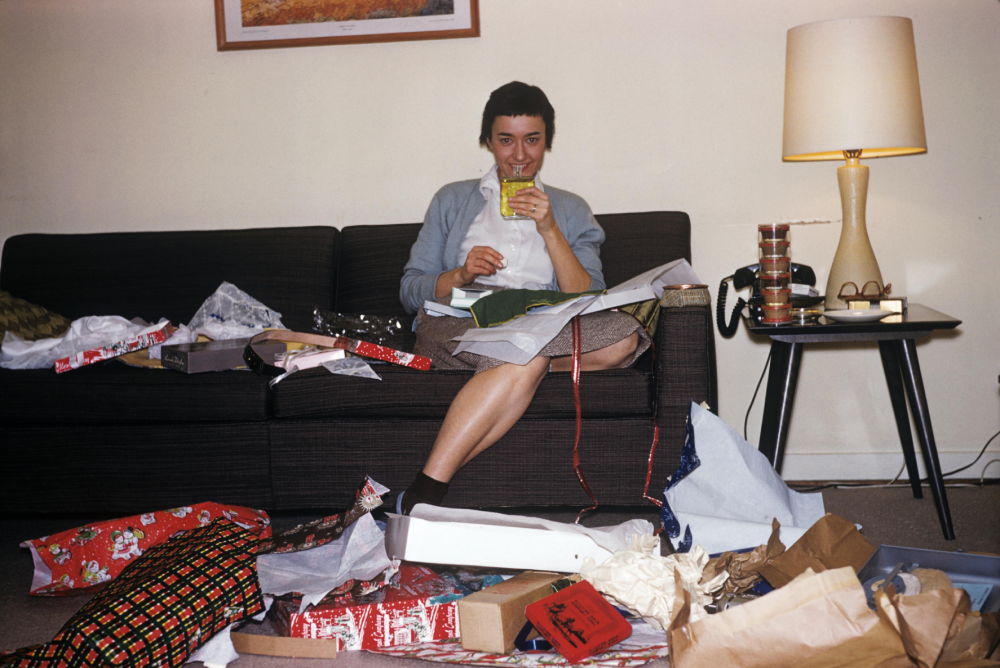 A woman sits on a sofa holding a glass, surrounded by opened Christmas presents and wrapping paper.